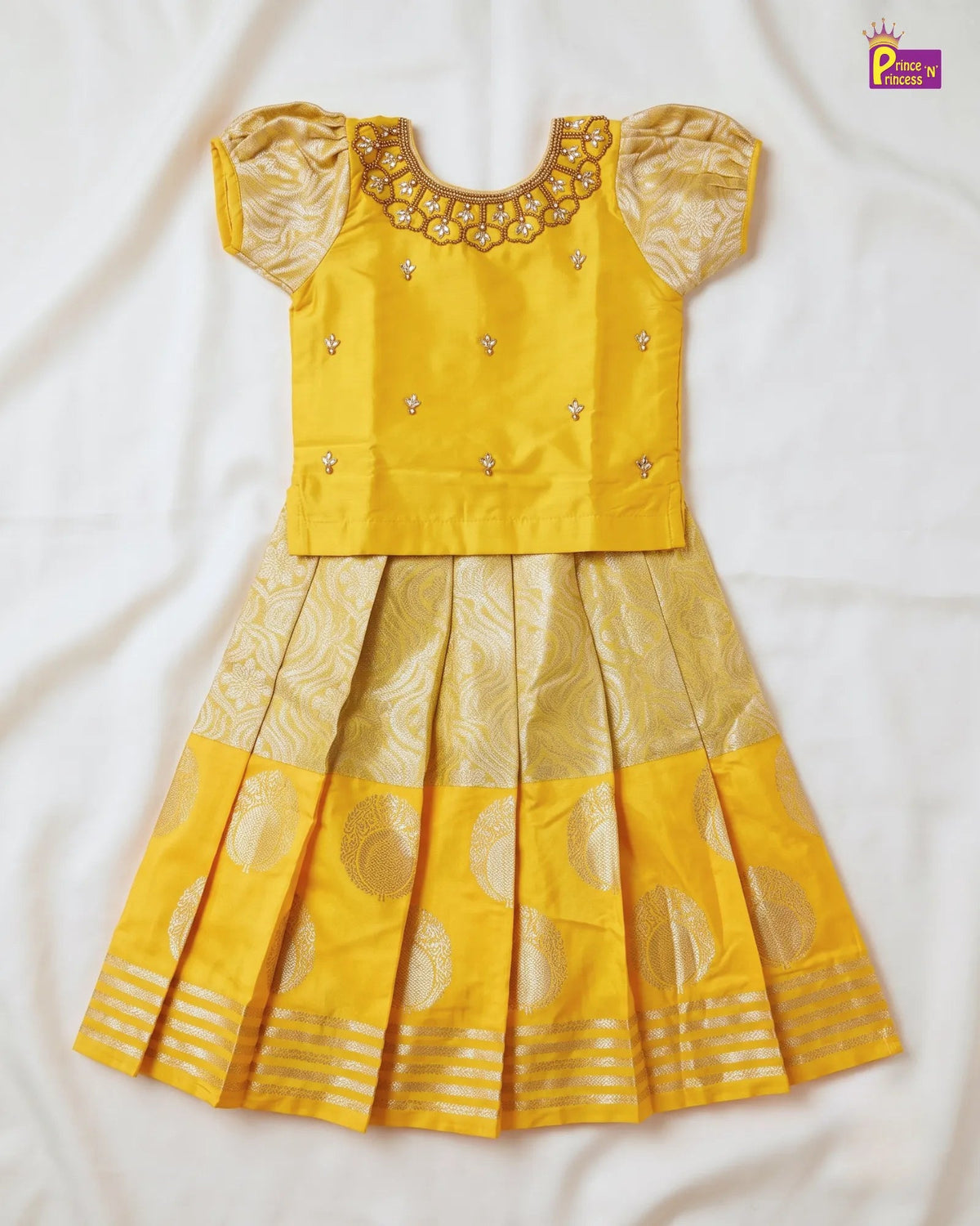 A yellow traditional Indian girl's dress set with a short-sleeved embroidered blouse and a pleated skirt featuring gold zari motifs, laid flat on a white fabric background.