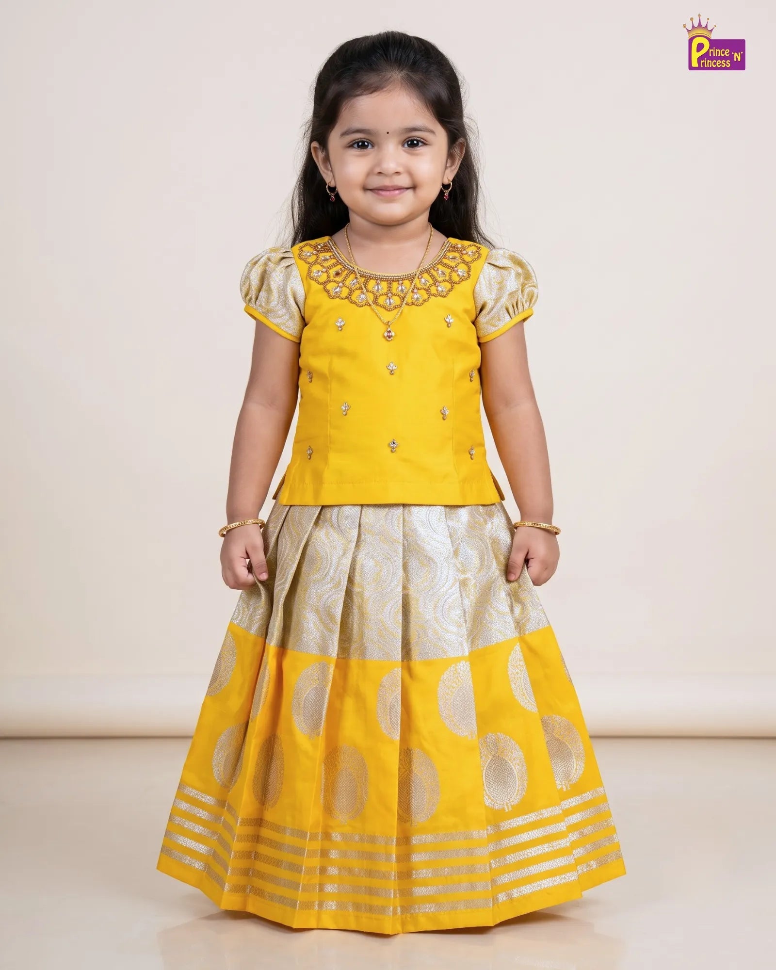 A young girl modeling a traditional Indian outfit consisting of a bright yellow embroidered kurta top and a matching pleated skirt with gold metallic motifs, set against a plain beige studio background.
