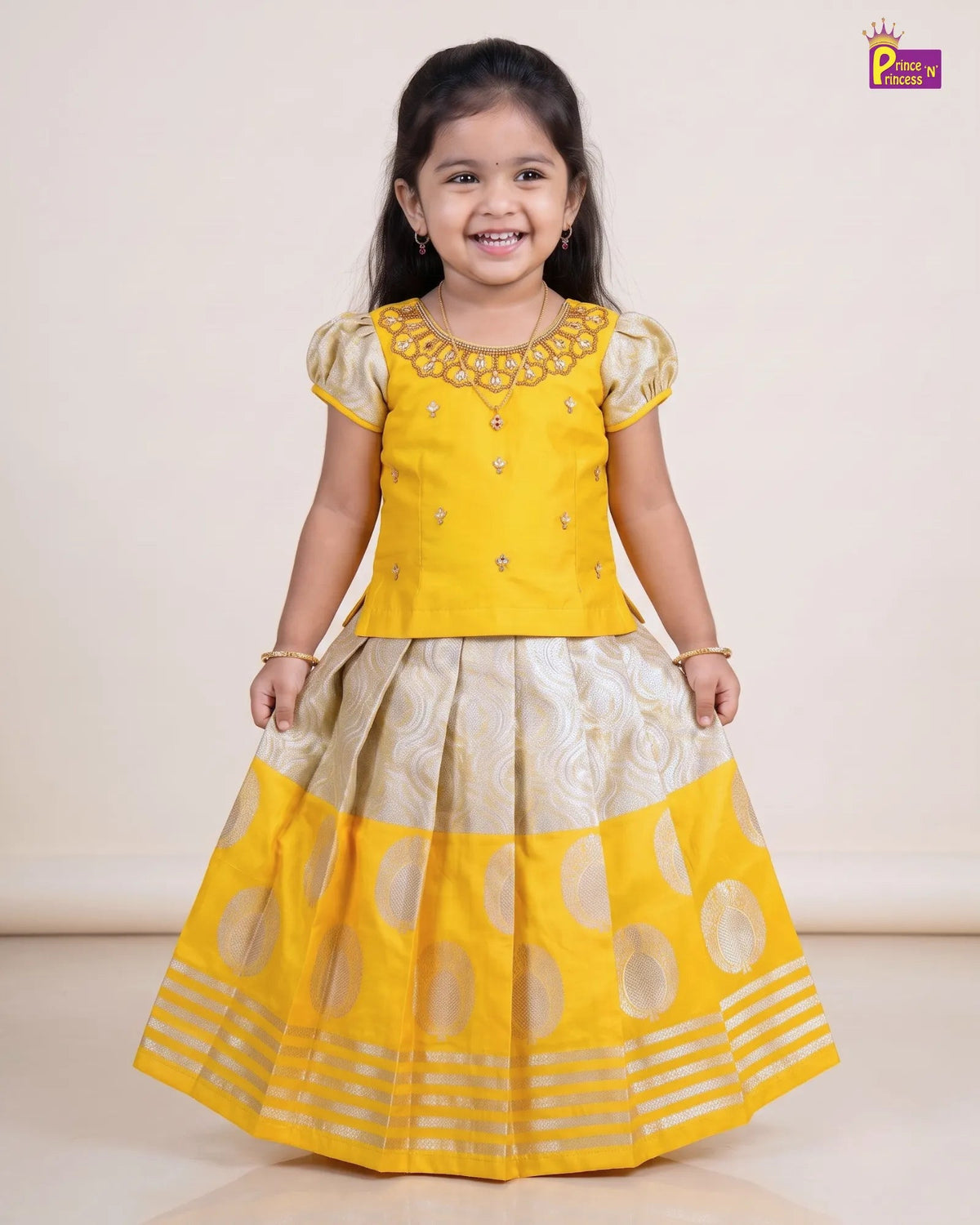 A young girl smiling and wearing a traditional Indian ethnic outfit consisting of a bright yellow embroidered kurta with short puff sleeves and a matching pleated skirt in yellow and beige with metallic gold patterns, set against a plain beige studio background.