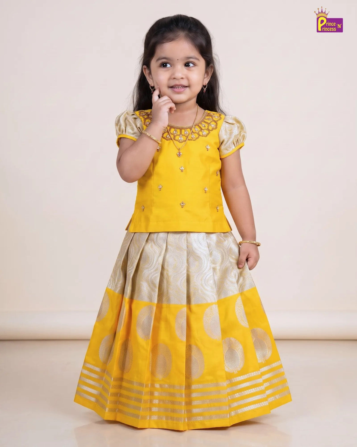 A young girl modeling a traditional Indian ethnic wear set consisting of a bright yellow embroidered kurta top and a matching pleated skirt with beige and yellow patterns, set against a plain beige studio background.