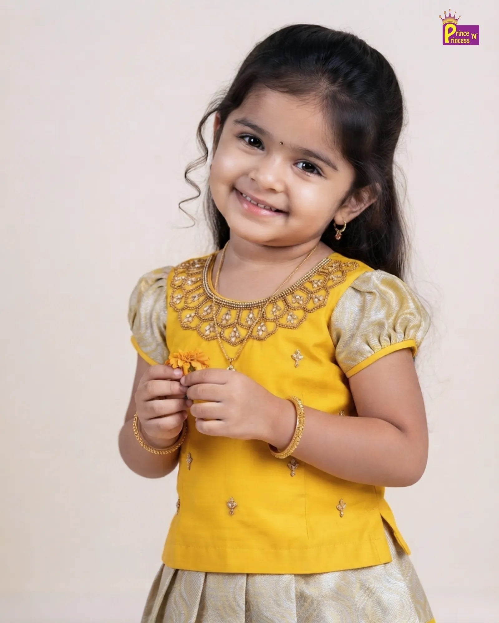 A young girl smiling and wearing a yellow traditional Indian outfit with gold embroidery and metallic gold sleeves, photographed against a plain light beige background.