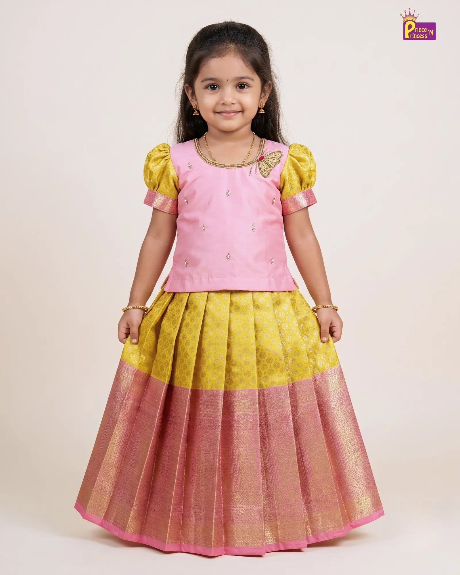 A young girl modeling a traditional Indian outfit consisting of a pink embroidered top with yellow puff sleeves and a matching pleated skirt in yellow and pink with a metallic border, set against a plain light beige background.
