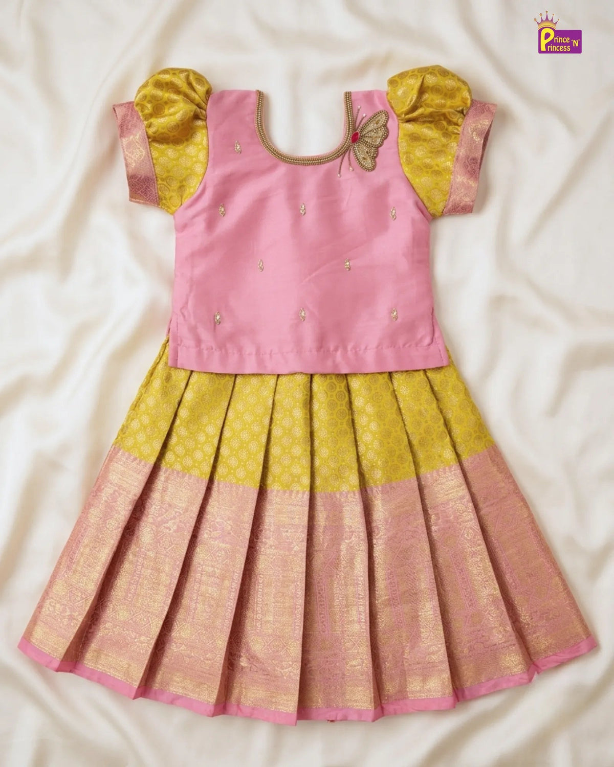 A traditional Indian girl's dress laid flat on a white satin fabric background. The dress features a pink sleeveless bodice with small gold embellishments, yellow puffed sleeves, and a pleated skirt transitioning from yellow to pink with a gold zari border.