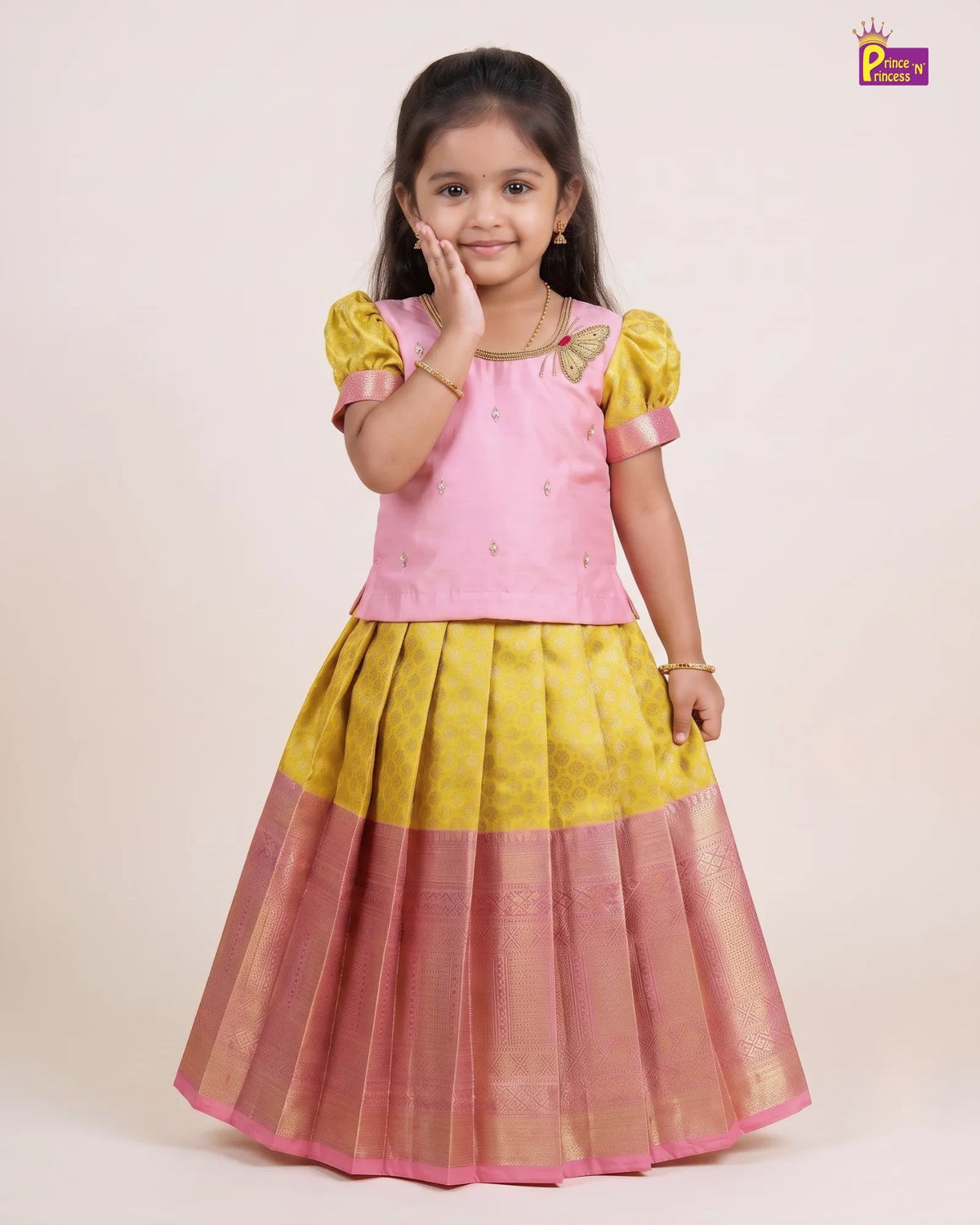 A young girl modeling a traditional Indian ethnic dress consisting of a pink top with yellow puffed sleeves and a pleated yellow and pink skirt with gold zari detailing, set against a plain beige studio background.