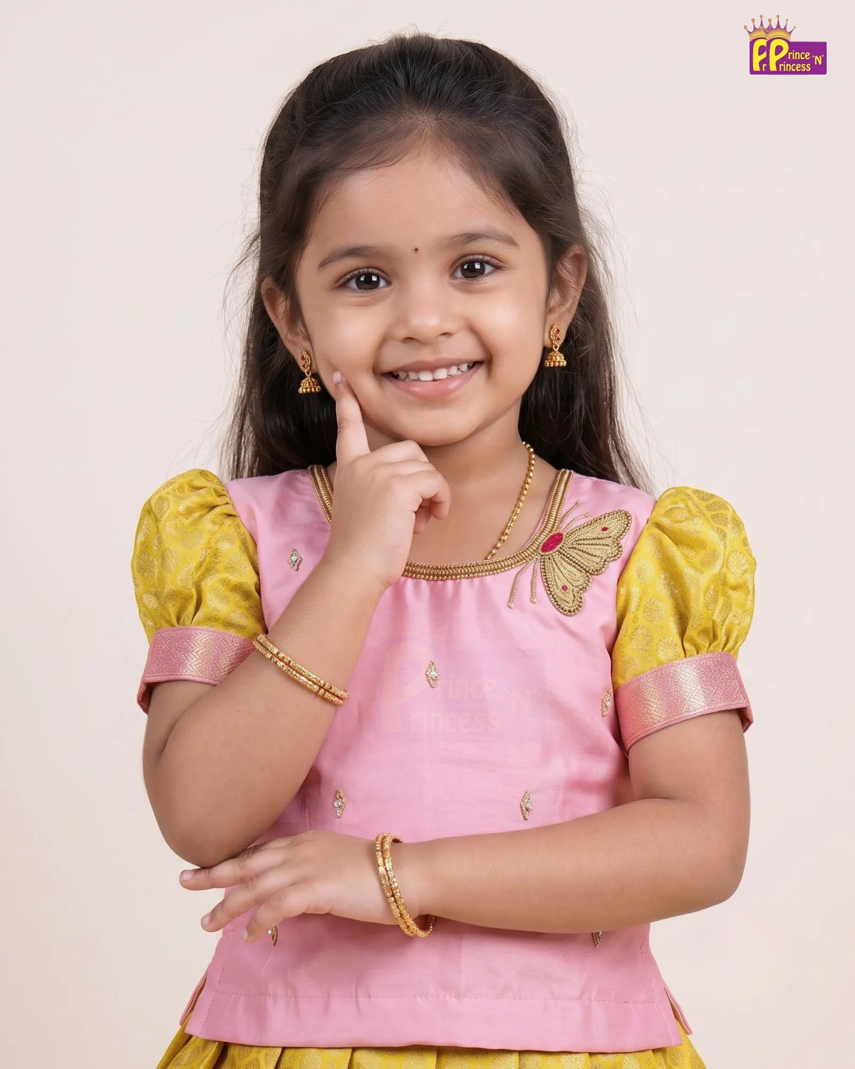 A young girl smiling and posing in a pink and yellow traditional Indian outfit with gold jewelry, set against a plain light beige background.