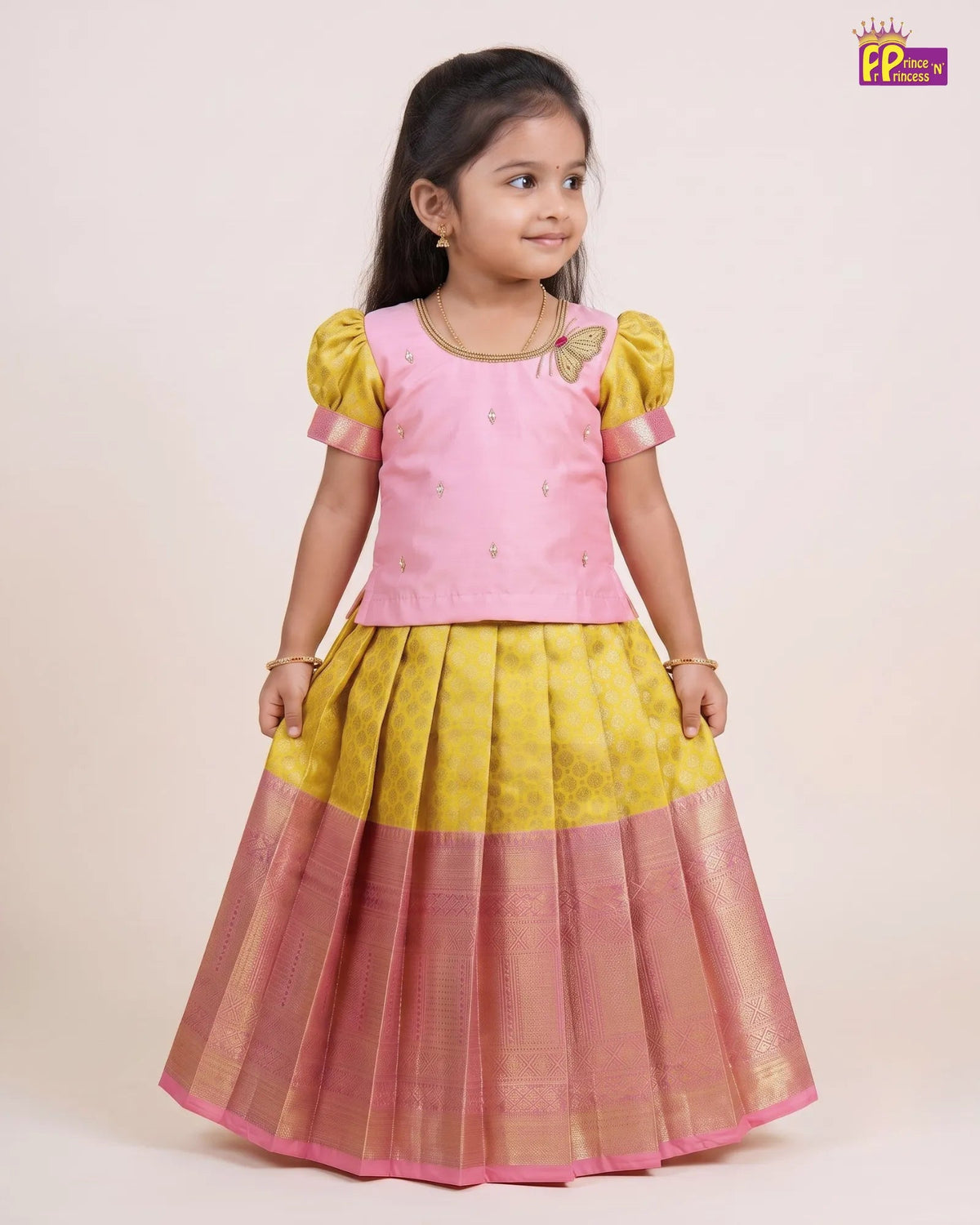 A young girl modeling a traditional Indian outfit consisting of a pink sleeveless top with yellow puff sleeves and a matching pleated skirt in yellow and pink with a gold zari border, set against a plain beige background.