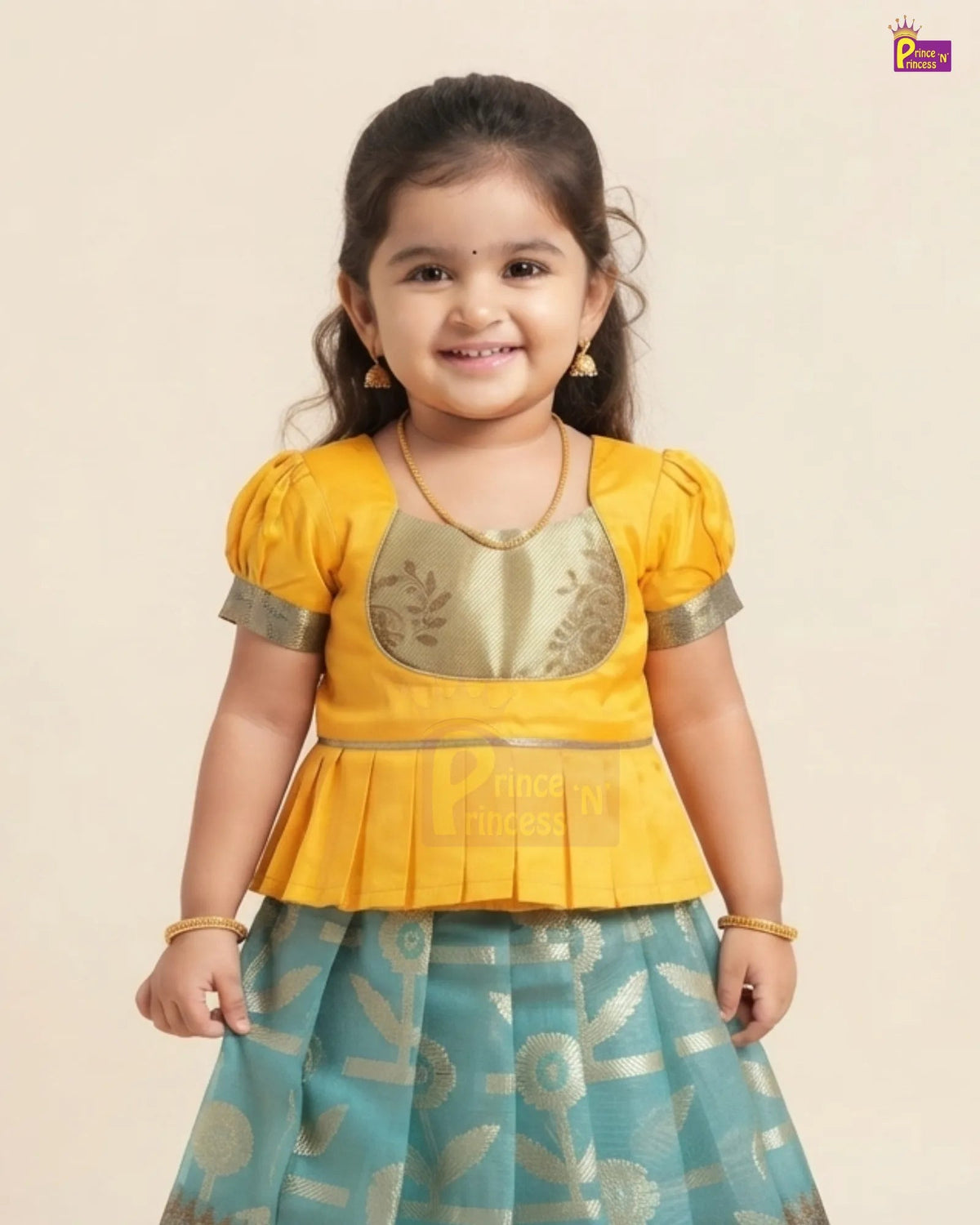 A young girl modeling a traditional Indian outfit consisting of a bright yellow peplum top with gold embroidery and a teal blue skirt with gold patterns, set against a plain beige background.