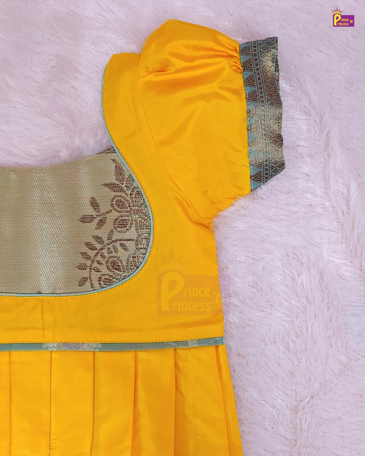 A close-up of a yellow children's dress or kurta laid flat on a soft pink faux-fur surface. The garment has a gold embroidered yoke, a light blue-green border trim, and a pleated skirt.