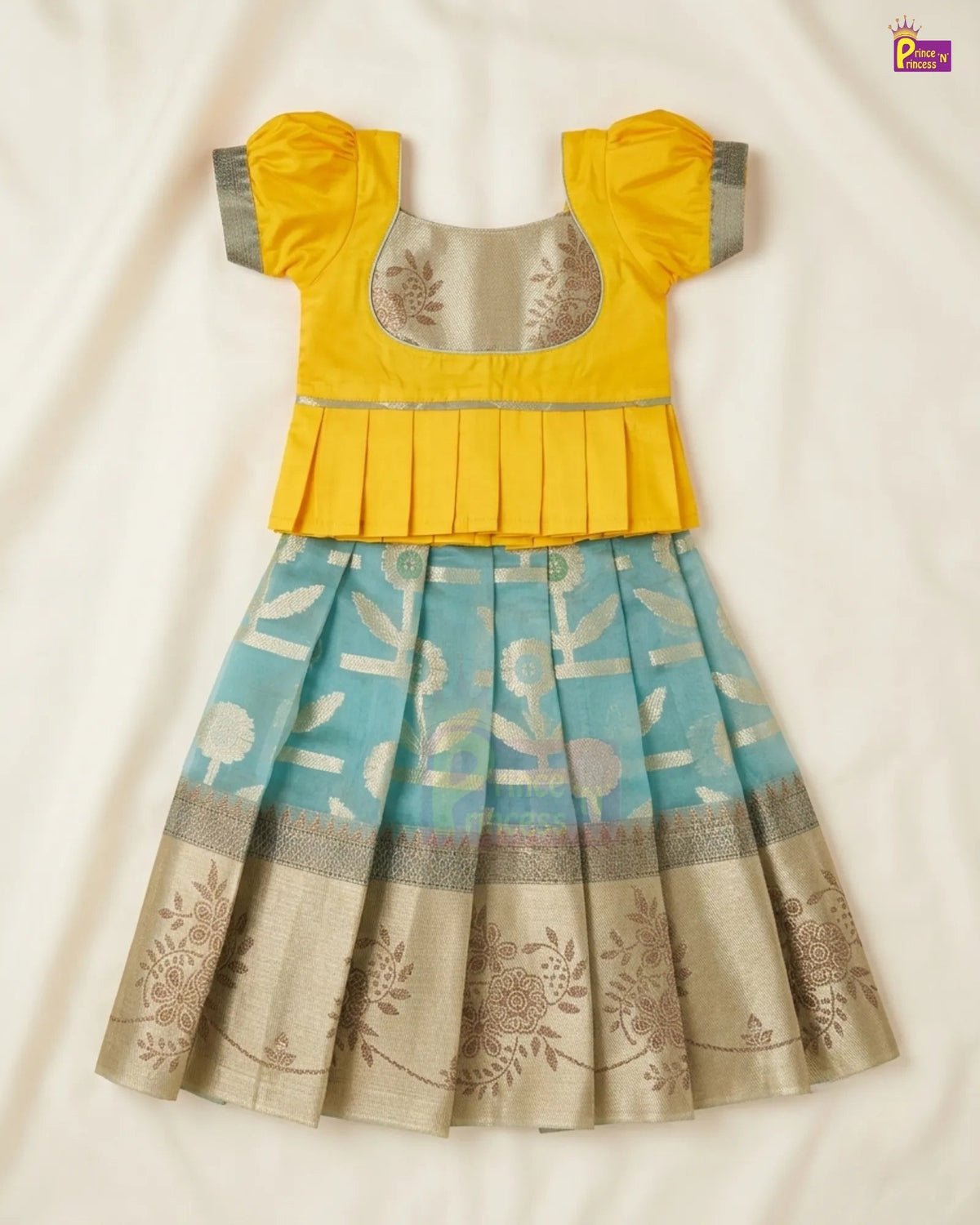 A traditional Indian girl's dress with a bright yellow short-sleeve top and a pleated light blue skirt featuring gold floral patterns and a beige border, laid flat on a cream fabric background.