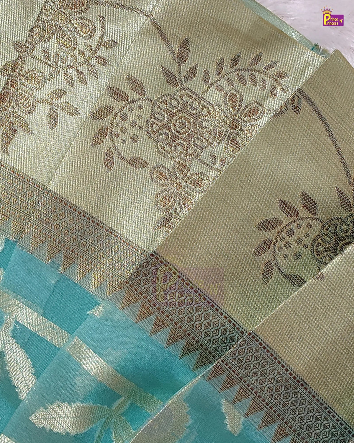 A close-up of a woven fabric, likely a saree, featuring a light beige body with brown floral and paisley zari motifs, a decorative border with geometric patterns, and a contrasting turquoise blue section with metallic silver leaf designs.